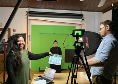 Photo of a woman holding a clapperboard in between a man with a camera, and a woman standing in front of a green screen.