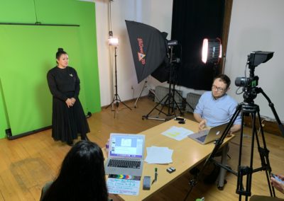 Photo of a woman standing in front of a green screen and camera. In front of her is a table with two people sitting with a laptop and papers.